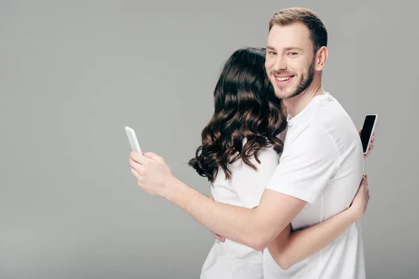 Pareja joven en camisetas blancas abrazándose mientras usa teléfonos inteligentes sobre fondo gris - foto de stock