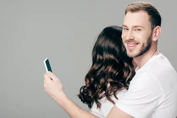 Красивий усміхнений чоловік обіймає жінку і використовує смартфон ізольовано на сірому — стокове фото