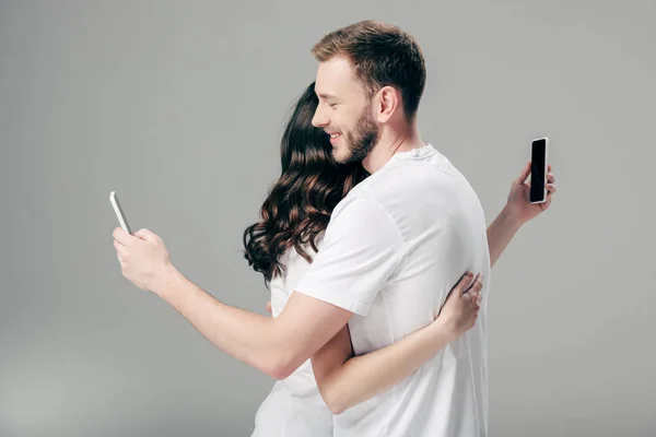 Young couple in white t-shirts hugging while using smartphones on grey background — Stock Photo