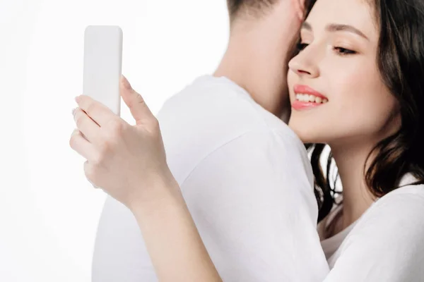 Atractiva chica sonriente usando teléfono inteligente mientras abraza novio aislado en blanco - foto de stock