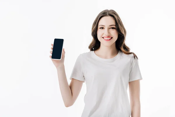 Hermosa chica alegre en camiseta blanca sosteniendo teléfono inteligente con pantalla en blanco y mirando a la cámara aislada en blanco - foto de stock
