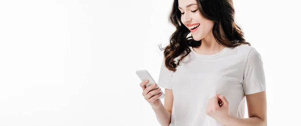 Панорамный снимок довольно веселой девушки в белой футболке с использованием смартфона, изолированного на белом — стоковое фото