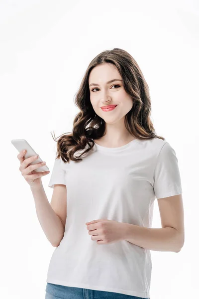 Красивая улыбающаяся девушка в белой футболке, держащая смартфон и глядя на камеру, изолированную на белом — стоковое фото
