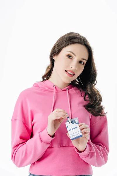 Atractiva chica sonriente en sudadera con capucha rosa conseguir cigarrillo de paquete con el logotipo de facebook aislado en blanco - foto de stock