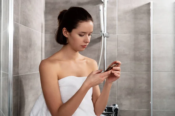 Jeune femme en serviette en utilisant smartphone dans la salle de bain — Photo de stock