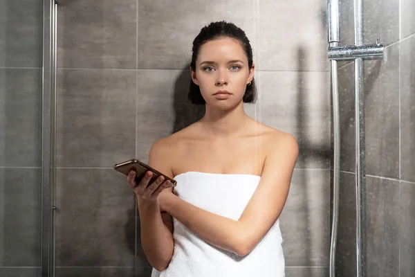 Front view of young woman in towel holding smartphone in bathroom — Stock Photo