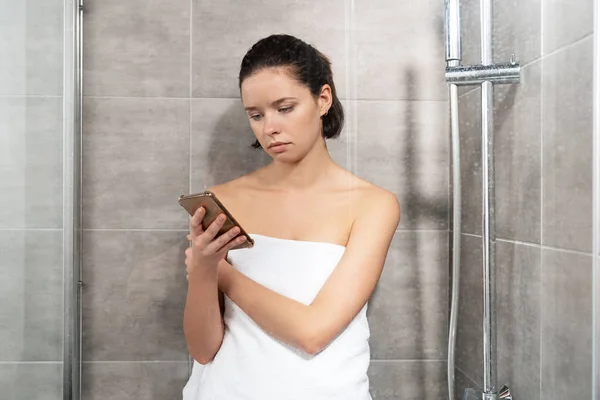 Pensive young woman in towel using smartphone in bathroom — Stock Photo