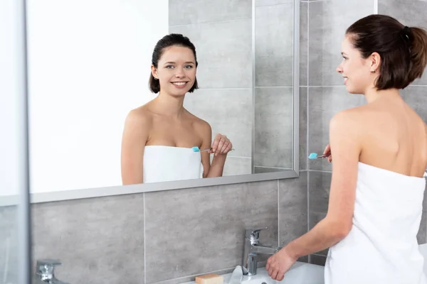 Симпатичная улыбающаяся девушка, держащая зубную щетку у зеркала в ванной комнате — стоковое фото