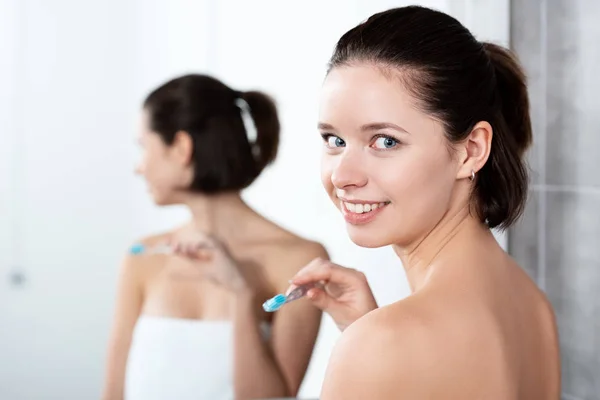 Bonita chica sonriente sosteniendo cepillo de dientes cerca del espejo en el baño - foto de stock