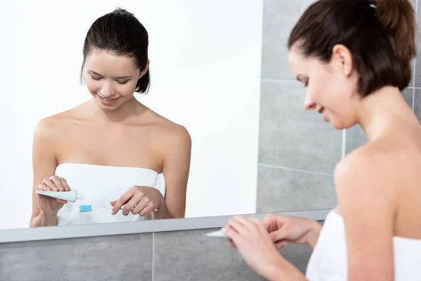 Jeune femme appliquant du dentifrice sur la brosse à dents près du miroir dans la salle de bain — Photo de stock