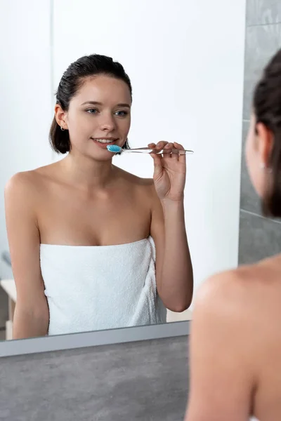 Веселая женщина в полотенце чистить зубы перед зеркалом — стоковое фото