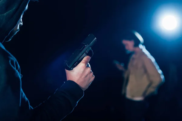 Vista recortada del asesino en la capucha con pistola y mirando a la mujer en negro - foto de stock