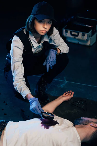 Investigator sitting near corpse and using magnifying glass at crime scene — Stock Photo