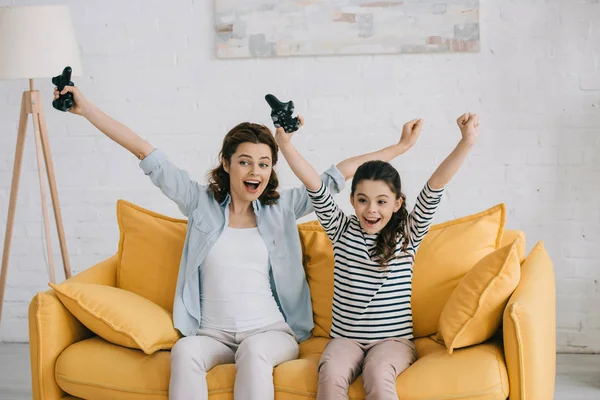KYIV, UCRANIA - 8 DE ABRIL DE 2019: Madre e hija emocionadas mostrando sí gesto mientras están sentadas en el sofá amarillo y sosteniendo joysticks - foto de stock