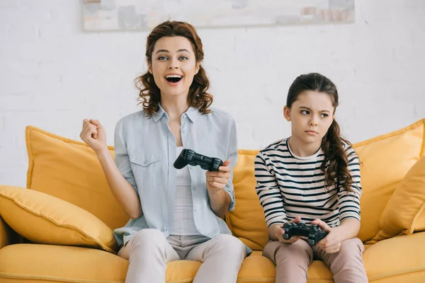 KYIV, UKRAINE - APRIL 8, 2019: Cheerful mother holding joystick and showing yes gesture while sitting near offended daughter — Stock Photo