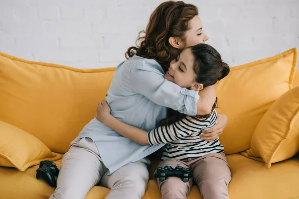 KYIV, UKRAINE - APRIL 8, 2019: Happy mother and daughter hugging while sitting with joysticks on sofa at home — Stock Photo