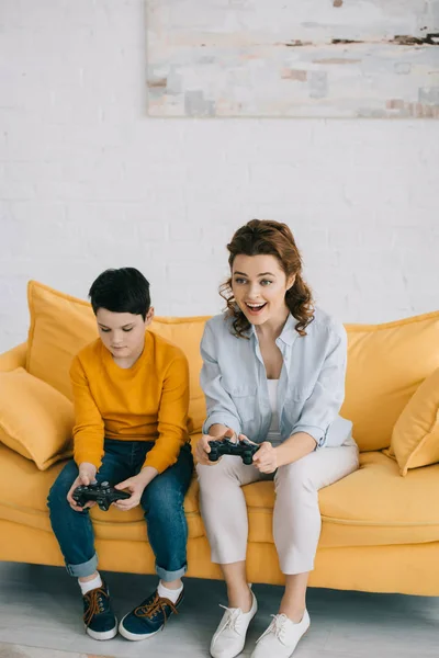 KYIV, UCRANIA - 8 de abril de 2019: Mujer emocionada jugando videojuegos mientras está sentada cerca de su hijo ofendido sosteniendo el joystick - foto de stock