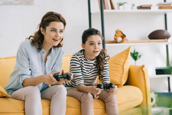 KYIV, UCRANIA - 8 DE ABRIL DE 2019: Madre e hija sonrientes jugando videojuegos mientras están sentadas en el sofá en casa - foto de stock