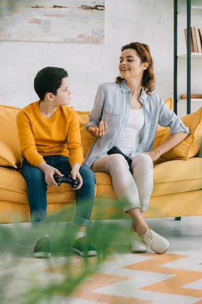 KYIV, UKRAINE - APRIL 8, 2019: Cheerful mother gesturing and looking at upset son holding joystick while sitting on sofa at home — Stock Photo