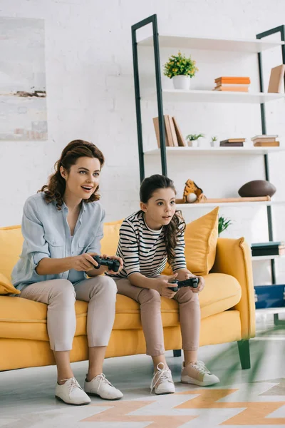 KYIV, UCRANIA - 8 DE ABRIL DE 2019: Madre e hija concentradas jugando videojuegos con joysticks mientras están sentadas en un sofá amarillo - foto de stock