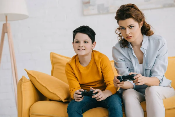 KYIV, UCRANIA - 8 DE ABRIL DE 2019: Niño sonriente y madre atenta jugando videojuegos con joysticks - foto de stock