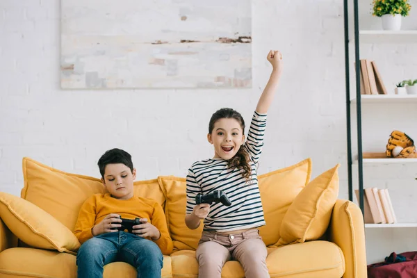 KYIV, UKRAINE - APRIL 8, 2019: Excited sister showing yes gesture while sitting near offended brother on yellow sofa — Stock Photo