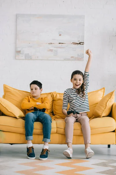 KYIV, UCRANIA - 8 DE ABRIL DE 2019: Hermana feliz sosteniendo el joystick y mostrando sí gesto mientras está sentada cerca de hermano molesto - foto de stock