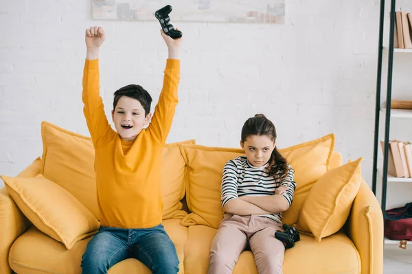 KYIV, UKRAINE - APRIL 8, 2019: Excited boy holding joystick in raised hands while sitting at yellow sofa near offended sister — Stock Photo