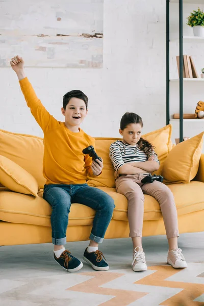 KYIV, UKRAINE - APRIL 8, 2019: Excited boy holding joystick and showing yes gesture while sitting near offended sister — Stock Photo