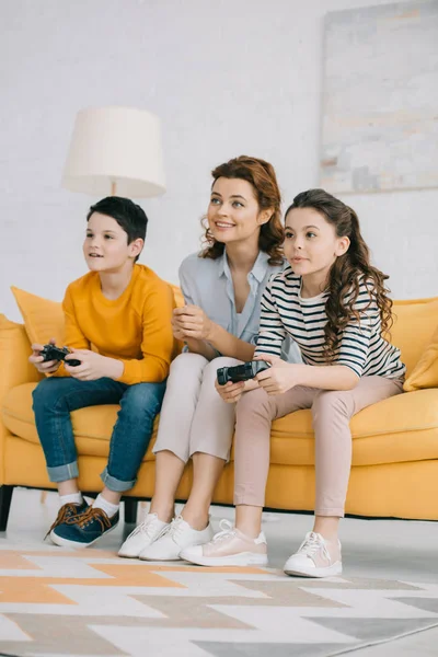 KYIV, UCRANIA - 8 DE ABRIL DE 2019: Feliz madre sentada en un sofá amarillo cerca de niños jugando videojuegos con joysticks - foto de stock