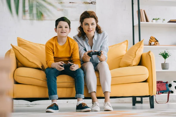 KYIV, UKRAINE - 8 de abril de 2019: Mãe e filho sorridentes jogando videogame com joysticks enquanto sentados no sofá amarelo — Stock Photo