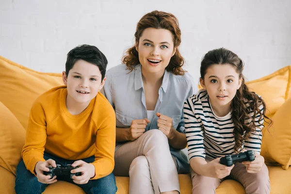 KYIV, UKRAINE - 8 AVRIL 2019 : Mère excitée assise sur un canapé près d'enfants jouant à un jeu vidéo avec des joysticks — Photo de stock