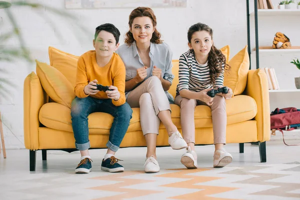 KYIV, UCRANIA - 8 DE ABRIL DE 2019: Mujer sonriente sentada en un sofá amarillo cerca de niños jugando videojuegos con joysticks - foto de stock