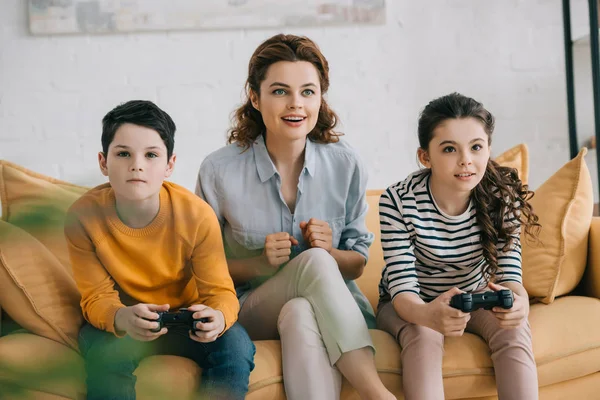 KYIV, UCRANIA - 8 de abril de 2019: Mujer alegre sentada en un sofá cerca de niños jugando videojuegos con joysticks - foto de stock