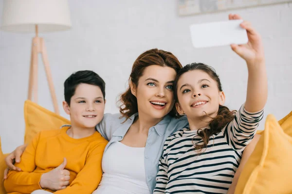 Fröhliches Kind macht Selfie lächelnd mit Mutter und Bruder, während es zu Hause auf dem Sofa sitzt — Stockfoto