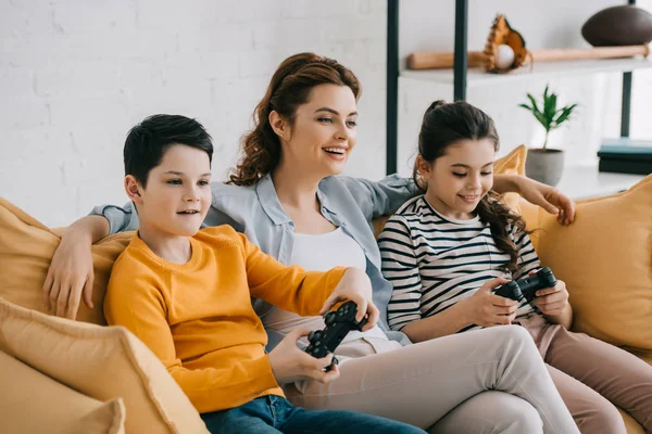 KYIV, UCRANIA - 8 DE ABRIL DE 2019: Mujer sonriente sentada en un sofá cerca de niños jugando videojuegos con joysticks - foto de stock