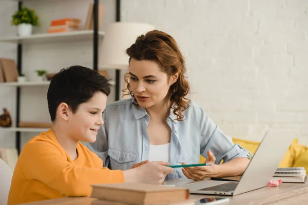 Lächelnde Mutter hilft entzückendem Sohn bei Hausaufgaben, während er am Schreibtisch mit Notizbuch sitzt — Stockfoto