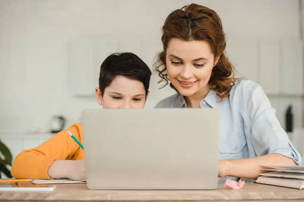 Mère heureuse avec fils mignon utilisant ordinateur portable ensemble tout en faisant des travaux scolaires à la maison — Photo de stock