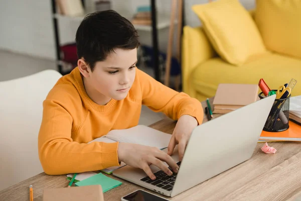 Niño atento usando el ordenador portátil mientras está sentado en el escritorio de madera y haciendo la tarea - foto de stock