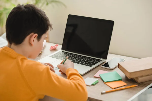 Schüler macht Hausaufgaben, während er am Schreibtisch neben Laptop mit leerem Bildschirm sitzt — Stockfoto