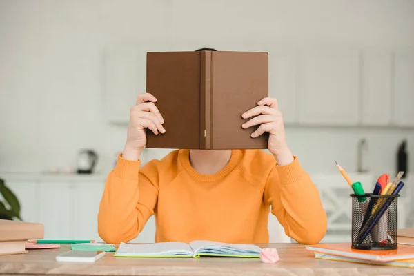 Niño ocultando la cara detrás de libro mientras está sentado en el escritorio y haciendo el trabajo escolar en casa - foto de stock