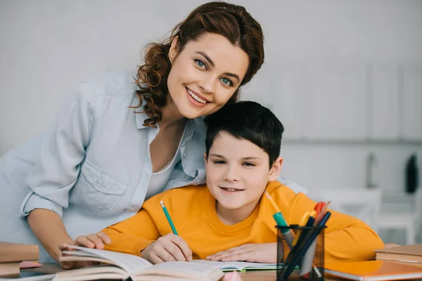 Feliz madre con adorable hijo sonriendo y mirando a la cámara mientras hacen los deberes juntos - foto de stock