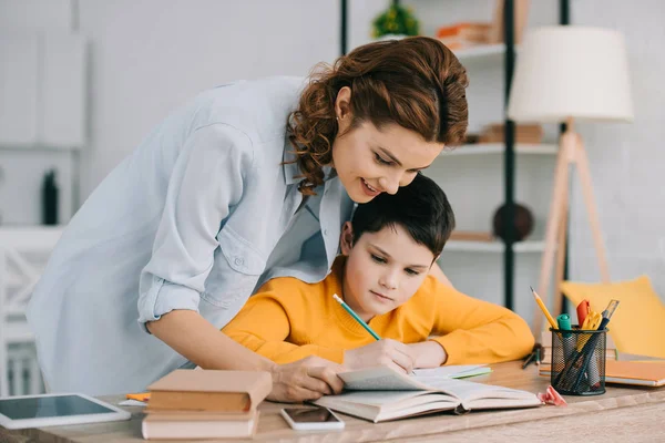 Jolie femme souriante aidant adorable fils faisant des travaux scolaires à la maison — Photo de stock