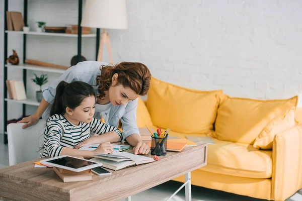 Hermosa mujer ayudando adorable hija haciendo trabajo escolar en casa - foto de stock