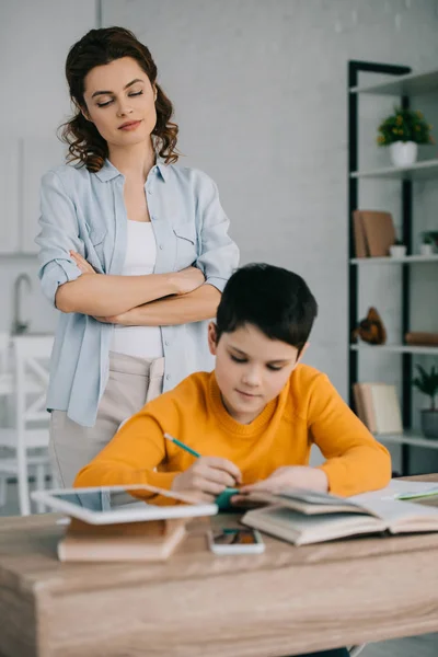 Foco seletivo de mãe atenta de pé com braço cruzado atrás do filho sentado na mesa e fazendo lição de casa — Fotografia de Stock