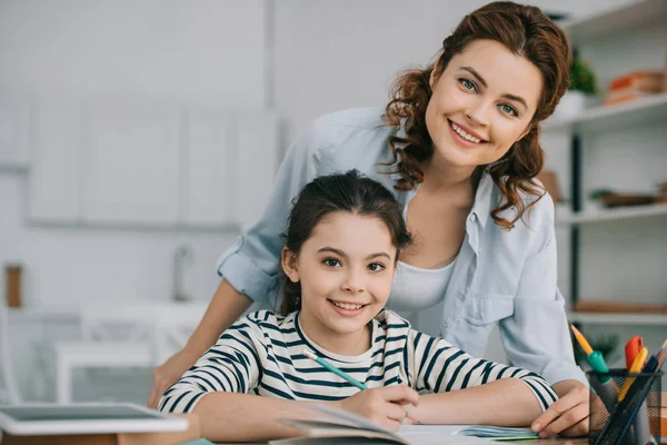 Happy mother with adorable daughter smiling at camera while doing schoolwork together at home — Stock Photo