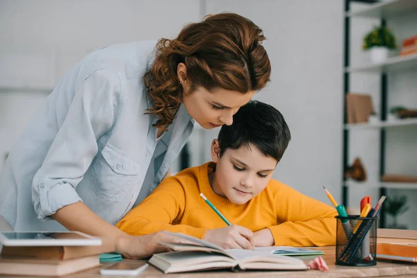 Schöne Frau, die in der Nähe von entzückendem Schreiben in Kopierbuch steht, während sie zu Hause Hausaufgaben macht — Stockfoto