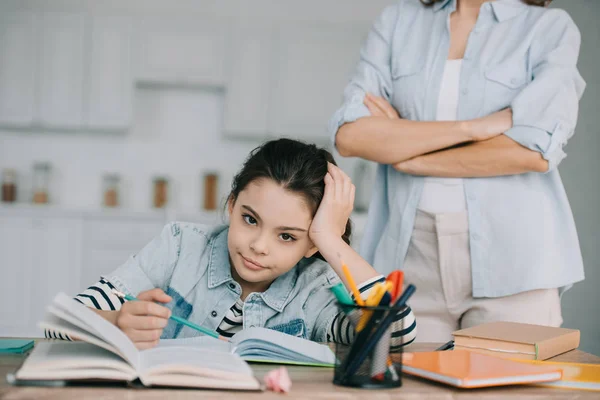 Vista parcial de la mujer de pie con los brazos cruzados detrás de hija aburrida haciendo la tarea - foto de stock