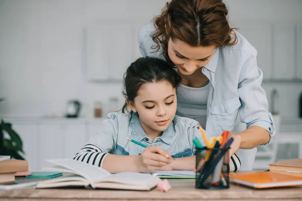 Adorable child writing in copy book while doing schoolwork near mother — Stock Photo