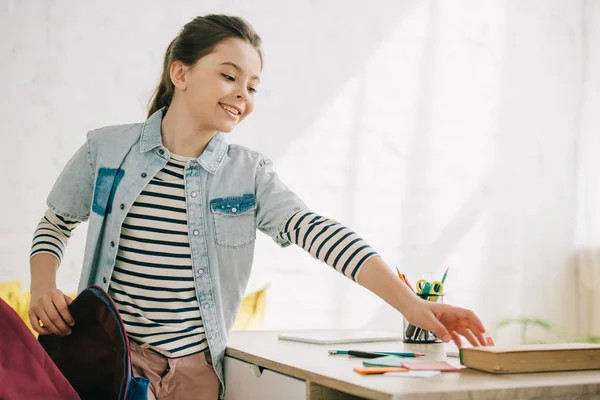 Smiling schoolchild packing back pack while standing near desk at home — Stock Photo